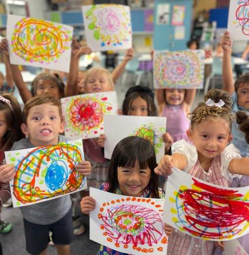 A group of young students are holding up their artwork and smiling. Their pages are a swirl of colors, dots, and squiggles.