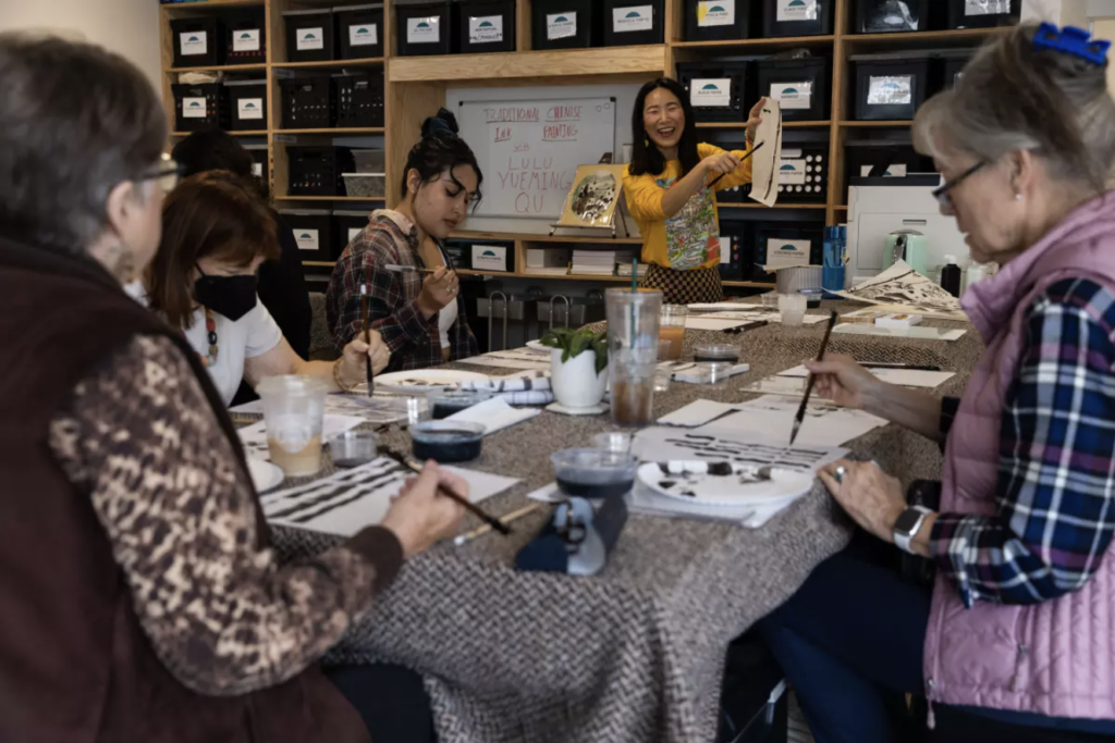 Five people are sitting at a table full of art supplies, painting. Lulu is wearing a yellow top and demonstrating what to do.